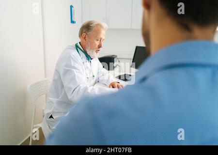 Nahaufnahme vom Rücken zum grauhaarigen Oberarzt, der einen medizinischen Mantel trägt und das Problem erklärt und ein Rezept am Tisch schreibt. Stockfoto