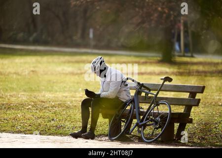 Stuttgart, Deutschland - 06. Januar 2022: Rennradfahrer machen auf der Parkbank eine Pause. Der männliche Athlet in Winterkleidung und Helm sitzt neben seinem Sport Stockfoto