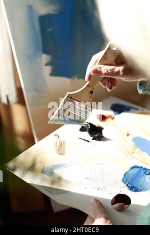 Die Hand einer älteren Frau mit Kunstpinsel, die Farbe aus der Palette in ihrem Studio bei natürlichem Licht aufgreift Stockfoto