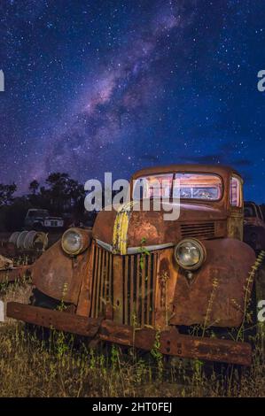 Ein alter, alter Ford-Pick-up, der in einem von Bäumen gesäumten grasbewachsenen Fahrerlager mit dem Milchstraßenkern im Hintergrund in der Nähe von Chillagoe, Queensland, in Australien, liegt. Stockfoto