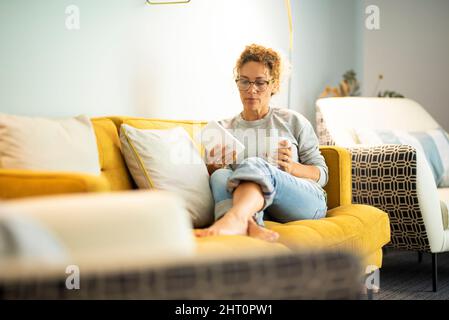 Selbstbewusste Erwachsene kaukasische Frau, die auf dem Sofa sitzt und ein Buch mit einem Tablet-Lesegerät für elektronische Geräte liest. Hübsche weibliche Menschen haben sich entspannt und genießen le Stockfoto