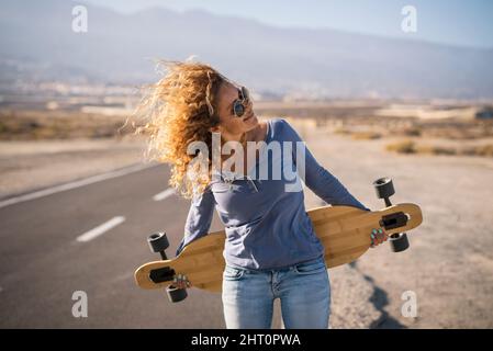 Hübsche Erwachsene Frau genießen das Leben und freien Lebensstil zu Fuß auf einer langen Asphaltstraße und halten lange Board Skate. Konzept der glücklichen schönen weiblichen Modder Stockfoto