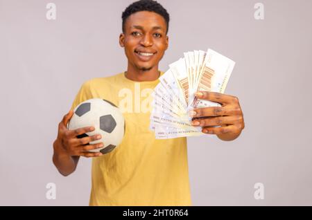 Sportwetten. Porträt Von Aufgeregt Black Guy Mit Fußball Und Geld Stehen Über Beige Hintergrund. Stockfoto