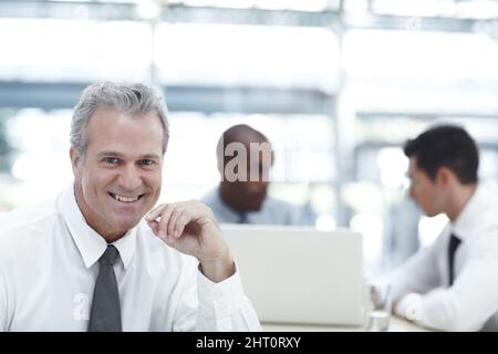 HES ein ausgewogener Arbeitgeber. Porträt eines reifen Geschäftsmannes, der lächelt, während er vor zwei jüngeren Kollegen sitzt. Stockfoto