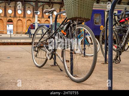 Ein Fahrrad mit einem Korb, der auf einer Bahnhofsplattform geparkt ist. Hinter dem Fahrrad befindet sich ein Zug und im Hintergrund eine Wand mit runden Fenstern. Stockfoto