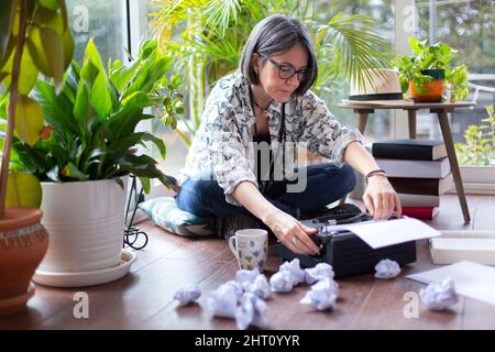 Erwachsene weibliche Schriftstellerin, die neben vielen zerknitterten Papieren ein neues leeres Blatt Papier auf die Schreibmaschine legte. Kreativblock. Stockfoto