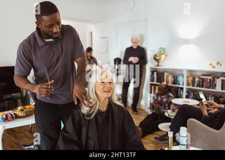 Männlicher Betreuer, der im Pflegeheim mit einer älteren Frau spricht, während er Haare schneidet Stockfoto