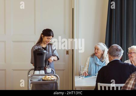 Lächelnde medizinische Mitarbeiterin, die älteren Frauen und Männern im Altersheim Kaffee serviert Stockfoto