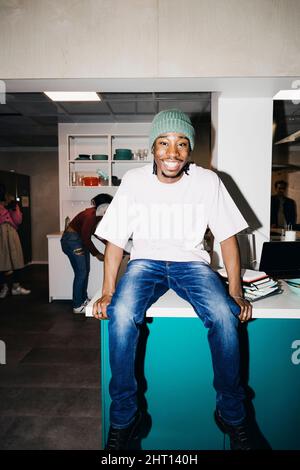 Porträt eines glücklichen jungen Mannes, der in der Küche im Studentenwohnheim auf dem Esstisch sitzt Stockfoto