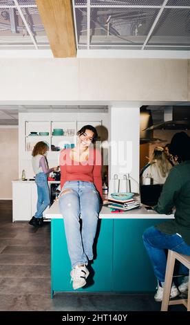 Porträt einer glücklichen jungen Frau in Casuals, die auf dem Esstisch im Studentenwohnheim sitzt Stockfoto