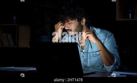 Überarbeiteter junger indischer Geschäftsmann, der spät am Arbeitsplatz eine Brille auszieht. Stockfoto