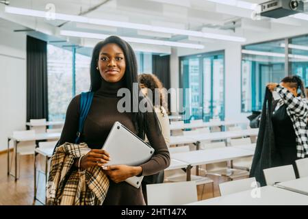 Porträt einer lächelnden jungen Frau mit Jacke und Laptop im Klassenzimmer Stockfoto