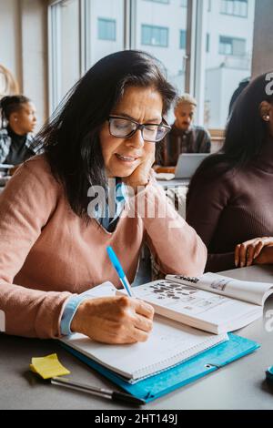 Reife Schülerin, die im Buch schreibt, während sie mit Freunden im Klassenzimmer sitzt Stockfoto