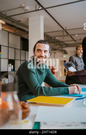 Lächelnder junger männlicher Student, der mit Büchern und Akten an einem Tisch in der Gemeinschaftsschule sitzt Stockfoto