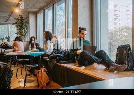Junger Mann mit Laptop sitzt in der Nähe von Freunden in der Community College Cafeteria Stockfoto