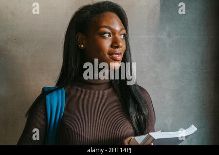 Nachdenkliche junge Frau, die das Buch gegen die graue Wand hält Stockfoto
