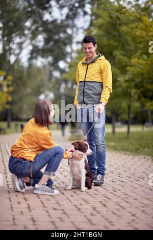 Junger kaukasischer Rüde, der mit seinem Hund an der Leine steht und das schöne Weibchen anschaut, lächelt, redet, unterhält sich Stockfoto