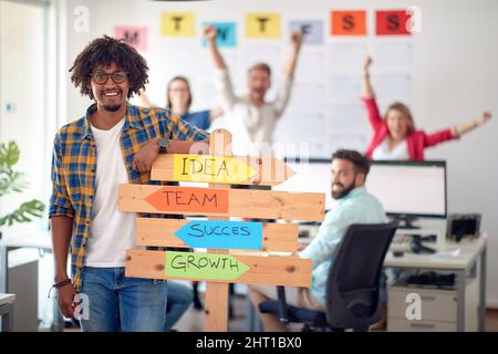 Ein junger Büroangestellter, der in einer angenehmen Atmosphäre im Büro für ein Foto posiert und gleichzeitig ein Schild mit Firmenslogans in der Hand hält. Mitarbeiter, Job, Büro Stockfoto