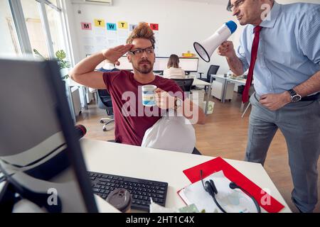 Ein junger männlicher Mitarbeiter in einer einheitlichen Atmosphäre im Büro ist arbeitsbereit, nachdem er von einem Manager aufgeweckt wurde, der ihn durch das Megaphon anschreit. Stockfoto