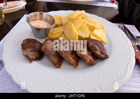 Gegrilltes Sirloin-Steak mit gerösteten, in Scheiben geschnittenen Kartoffeln, alles auf einem weißen Teller. Eine Schüssel mit heißer Sauce. Spanisches Speisekonzept. Stockfoto