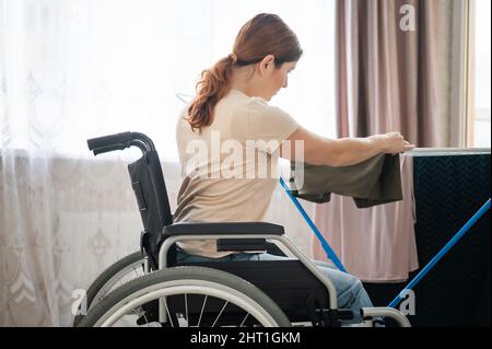 Kaukasische Frau im Rollstuhl hängt nasse Wäsche an einem Trockner. Stockfoto