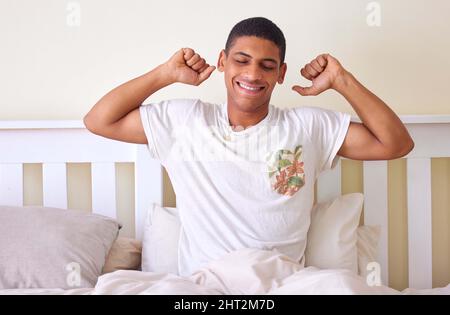 Ich liebe dieses erfrischte Gefühl am Morgen. Ausgeschnittene Aufnahme eines hübschen jungen Mannes, der in den frühen Morgenstunden im Bett sitzt und sich ausdehnt Stockfoto