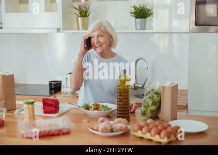 Glückliche, gealterte Frau, die in Innenräumen mit dem Mobiltelefon spricht Stockfoto