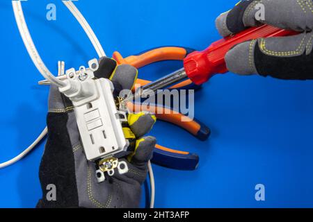 Ein Elektriker hält eine Steckdose mit der Hand in einem Handschuh und fixiert eine Schraube mit einem Schraubendreher Stockfoto