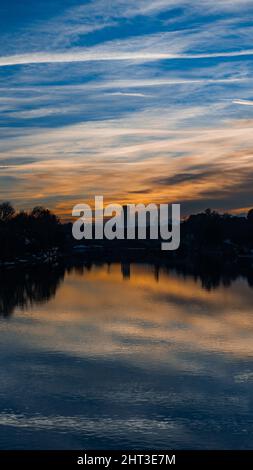 Vertikale Aufnahme von faszinierendem Seewasser, das in der Nähe von Parkbäumen reflektiert wird, und einem bewölkten Himmel bei Sonnenuntergang Stockfoto