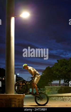 Schlafen Sie nie - fahren Sie einfach. Aufnahme eines Mannes, der nachts unter einer Straßenbeleuchtung Tricks auf seinem Fahrrad macht. Stockfoto