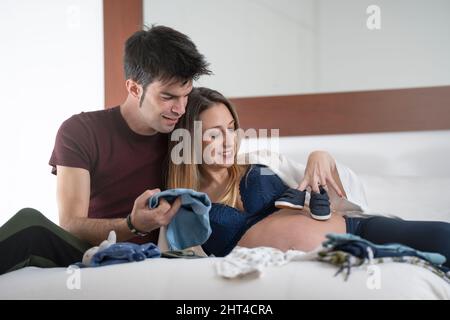 Junge Schwangerin mit ihrem Partner, der mit Babykleidung im Bett spielt Stockfoto
