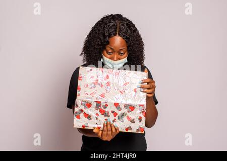 Junge schwarze Frau mit Gesichtsmaske sieht in einer Geschenkbox aus Stockfoto