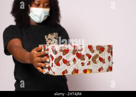 Eine junge afrikanische Frau, die eine Gesichtsmaske trägt und ein Geschenk ausdehnt, als ob sie es jemandem gibt Stockfoto