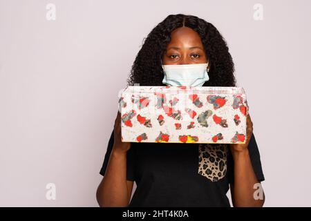 Junge schwarze Frau trägt eine Gesichtsmaske und hält ein Geschenk Stockfoto