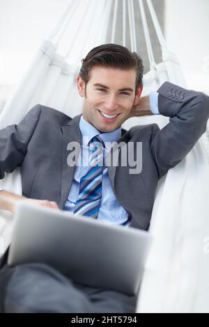 Entspannendes Business-Abenteuer. Ein junger Geschäftsmann liegt auf einer Hängematte, während er an seinem Laptop arbeitet. Stockfoto
