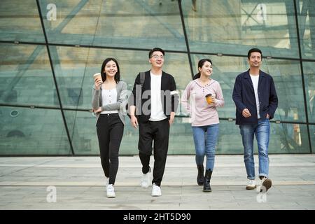 Eine Gruppe von vier jungen asiatischen Menschen, die sich unterhalten und auf der Straße in der modernen Stadt fröhlich und lächelnd spazieren gehen Stockfoto