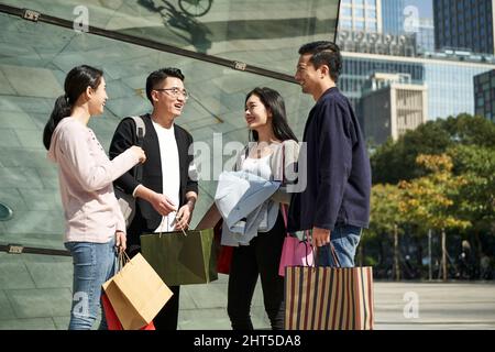 Eine Gruppe von vier jungen asiatischen Menschen, die auf der Straße stehen und sich unterhalten, unterhalten sich während eines Einkaufsbummel glücklich und lächelnd Stockfoto