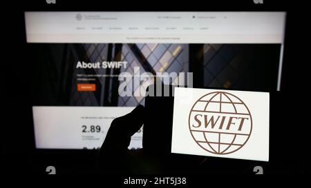 Person, die Mobiltelefon mit dem Logo des Banktransaktionssystems SWIFT auf dem Bildschirm vor der Webseite hält. Konzentrieren Sie sich auf die Telefonanzeige. Stockfoto