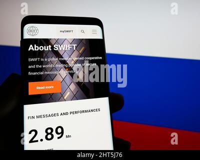 Person, die Mobiltelefon mit der Website des Banktransaktionssystems SWIFT auf dem Bildschirm vor der russischen Flagge hält. Konzentrieren Sie sich auf die Telefonanzeige. Stockfoto