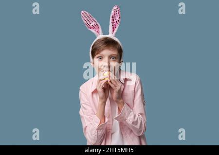 Schöner junger Mann in beiläufiger Kleidung rosa Hemd und Hase osterohren macht verschiedene Gesten mit den Handflächen seiner Hände mit einem Ei in der Hand, sieht sehr lustig und lustig osterhase Konzept. Stockfoto