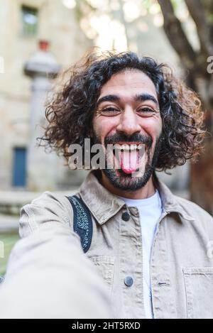 Entzückter bärtiger Mann, der Zunge zeigt und die Kamera anschaut, während er Selbstporträt macht und Spaß auf der Straße der Stadt hat Stockfoto