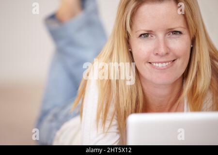 Online ein Buch lesen. Eine schöne reife Frau, die auf ihrem Laptop arbeitet, während sie auf dem Boden liegt. Stockfoto