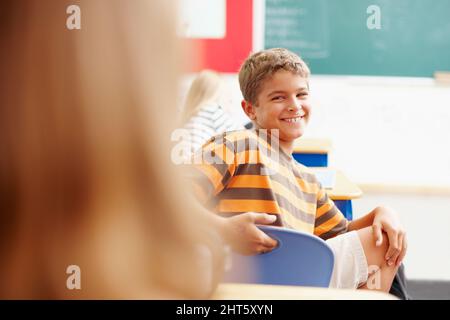 Holen Sie sich die Aufmerksamkeit des coolen Kindes in der Klasse. Lächelnder Junge dreht sich in der Klasse um, um einen Klassenkameraden zu betrachten - Copyspace. Stockfoto