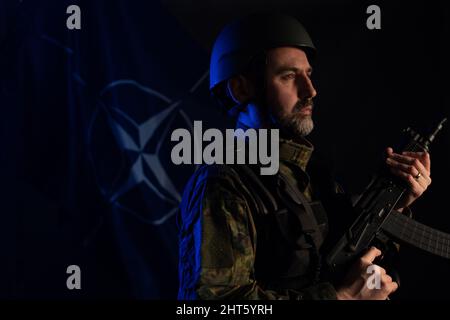 Soldat in Militäruniform und Helm mit Waffe mit NATO-Flagge im Hintergrund. Stockfoto