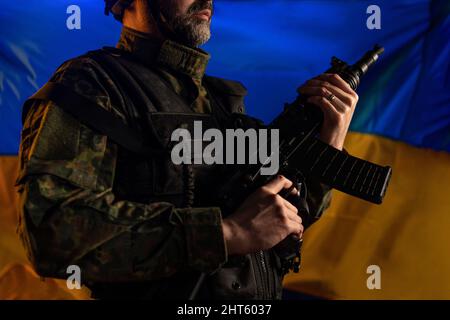 Nahaufnahme eines Soldaten in Militäruniform mit Waffe und ukrainischer Flagge im Hintergrund. Stockfoto