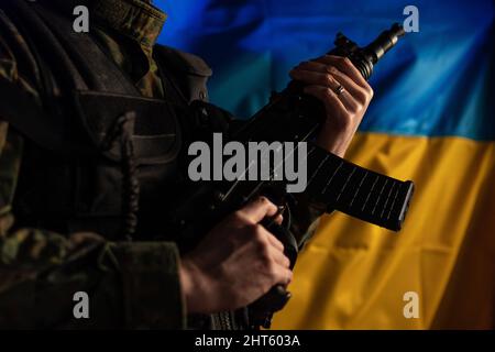 Nahaufnahme eines Soldaten in Militäruniform mit Waffe und ukrainischer Flagge im Hintergrund. Stockfoto