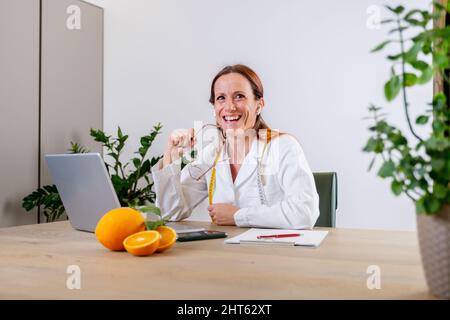 Porträt einer lächelnden jungen Ernährungswissenschaftlerin in ihrem Büro in einem medizinischen Kleid mit frischen Orangen. Dietologe macht einen Diätplan Stockfoto