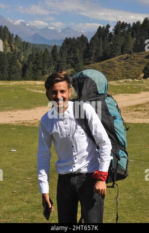 Vorderansicht eines glücklichen jungen Mannes, der die Kamera ansieht und einen Fallschirmrucksack im Berg trägt Stockfoto