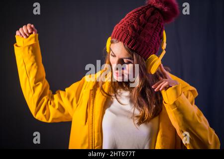 Porträt einer jungen brünette kaukasischen Frau mit Kopfhörern und gelber Jacke auf schwarzem Hintergrund Stockfoto
