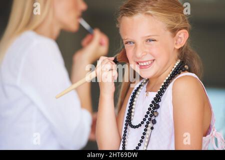 Make-up wie Mama. Das kleine Mädchen versucht, sich das Make-up ihrer Mutter anzuprobieren, während ihre Mutter im Hintergrund ist. Stockfoto